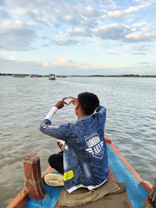 A person in a denim jacket sits on a blue boat, making a heart shape with their hands while looking out over a calm body of water at sunset.