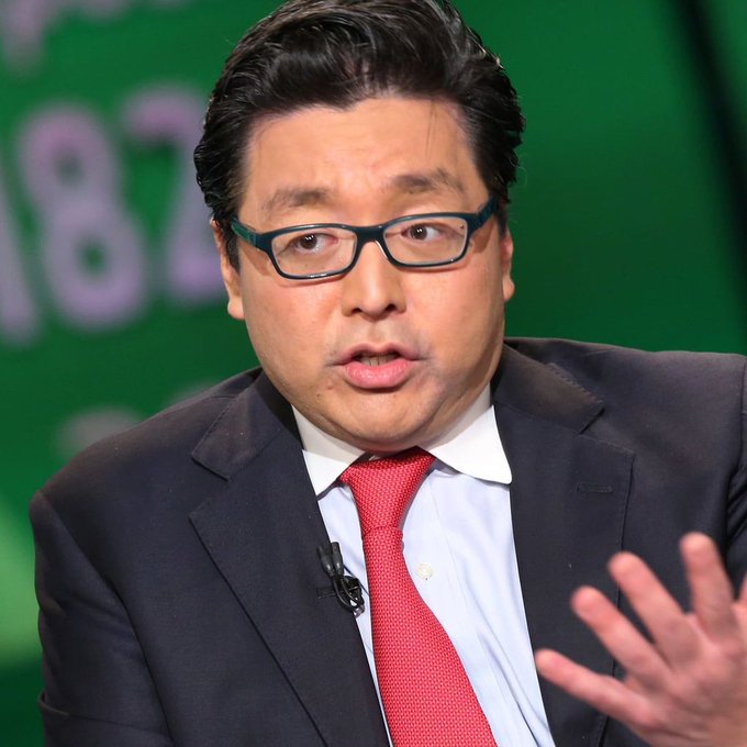 A man wearing a dark suit, red tie, and green-rimmed glasses speaks while gesturing with his hand against a green background.