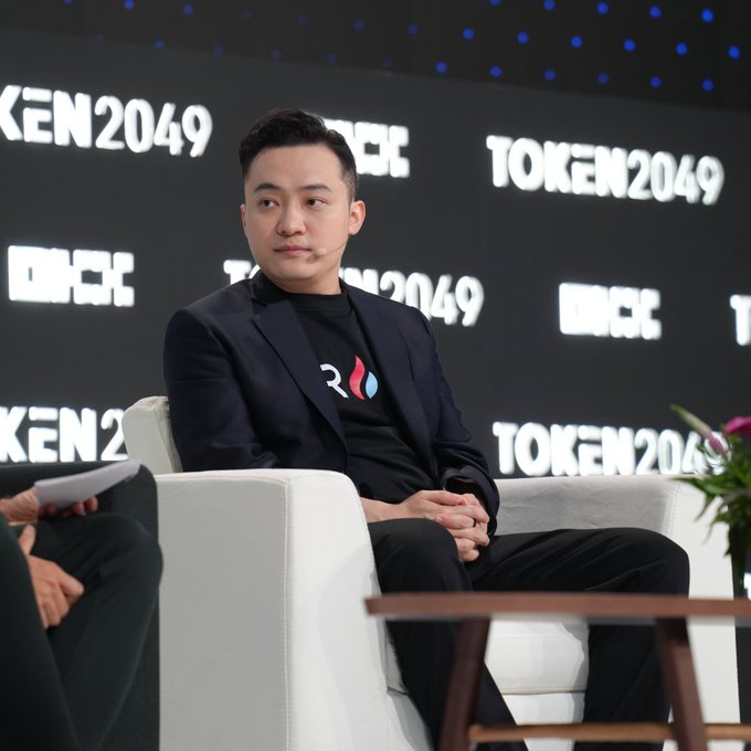 A man in a black suit seated at the TOKEN2049 conference with a '2049' branded black t-shirt.