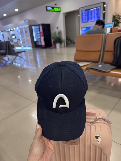 A hand holds up a navy blue baseball cap with a white logo in front of a pink suitcase at what appears to be an airport terminal.