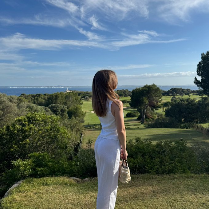 A woman in a white outfit stands overlooking a lush green golf course with the ocean and blue sky in the distance, embracing a serene start to "happy monday!"