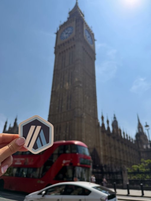 A photo of Big Ben in London with a red double-decker bus in the background, while a hand in the foreground holds a hexagonal sticker with a blue and white logo.