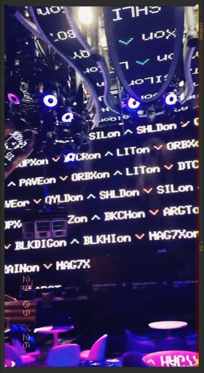 A dark, mirrored photograph of a stock market trading floor featuring electronic boards displaying stock tickers such as 'CHILI', 'UOX', 'LITon', 'SHLD', and 'ORBX'.