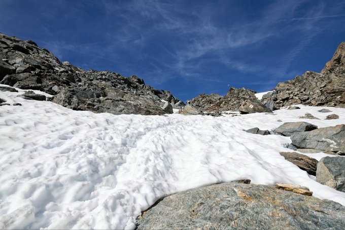 A landscape photo of a snowy mountain slope with rocky outcrops under a blue sky with wispy clouds.