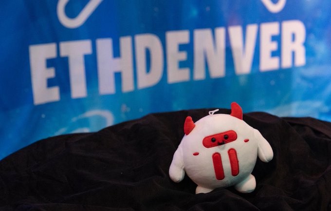 A small white plush toy with red devil horns and a red devil face stands on dark fabric, with a blue banner reading "ETHDENVER" in the background.