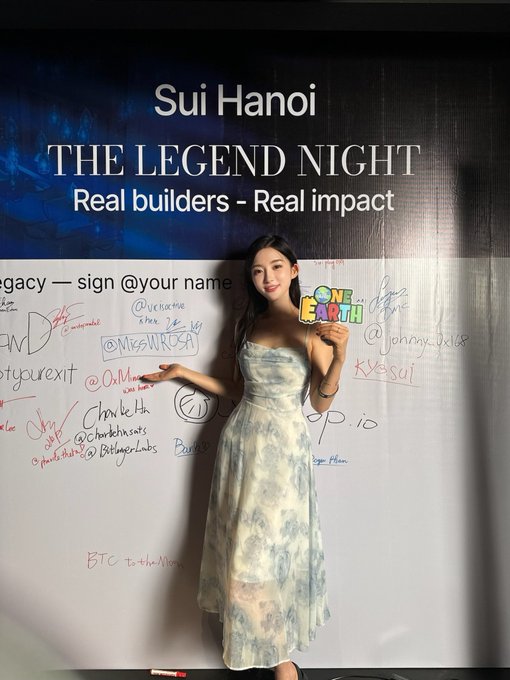 A woman in a light blue floral strapless dress poses in front of a signature wall displaying the text "Sui Hanoi - THE LEGEND NIGHT - Real builders - Real impact" along with various handwritten signatures and social media handles.