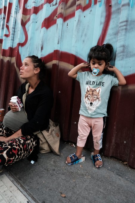 A woman holding a coffee sits next to a young child wearing a 'WILD LIFE' wolf-print t-shirt and a pacifier, standing against a graffiti-covered metal wall.