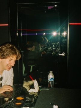 A candid photo of a smiling young person in a white t-shirt holding a gaming controller, seated in a dark venue with red LED strip lighting.