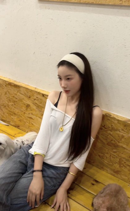 A young woman with long dark hair wearing a white off-shoulder top and a white headband sits against a wooden paneled wall, with a small white dog beside her and what appears to be a cat in the foreground. Her relaxed, contemplative pose and soft expression seem to mirror the hopeful yet uncertain tone of the post about checking back on a meme coin (@xdog_meme) and hoping to "break even," as she gazes off thoughtfully.