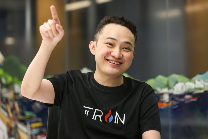 A smiling man in a black TRON-branded t-shirt points upward, standing in what appears to be an office with a cityscape mural in the background.