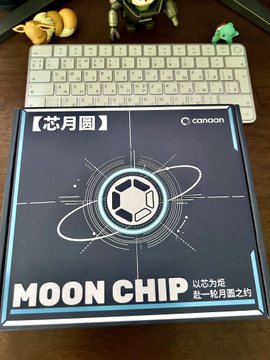 A Canon product box for 'MOON CHIP' (ムーンチップ), labeled '芯月層' and 'いなか月 壱〜拾月届之約', sitting on a desk next to a white Apple keyboard with small figurines.