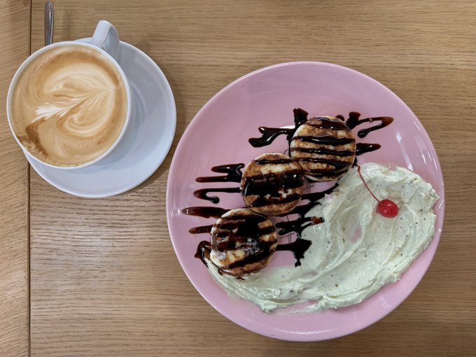 A cup of cappuccino with latte art sits next to a pink plate containing three small pancake stacks drizzled with chocolate syrup, whipped cream, and a cherry on a wooden table.