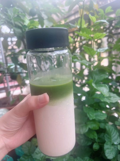 A hand holds a clear glass bottle with a black lid containing a layered matcha latte drink, featuring a green top layer and a creamy white bottom layer, set against a background of lush green plants.
