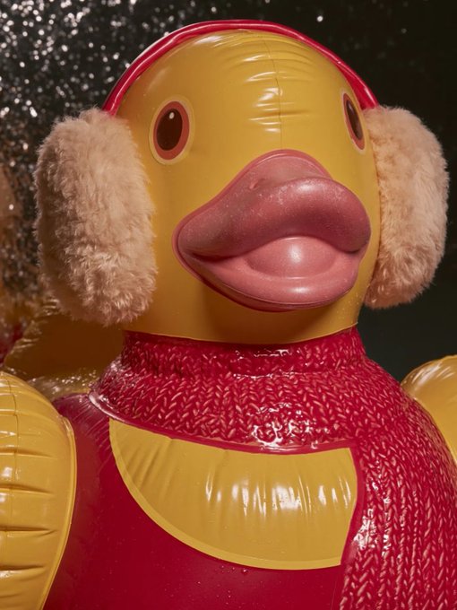 A close-up of an inflatable yellow duck wearing pink earmuffs, a red knitted sweater, and featuring prominently large pink lips.