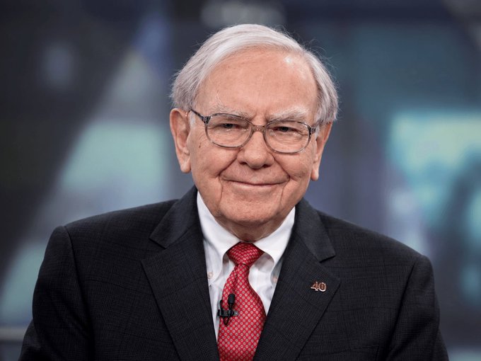Warren Buffett, wearing a dark suit, red patterned tie, and glasses, smiles slightly in what appears to be a television studio setting.