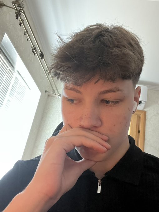 A young man with brown curly hair wearing a black zip-up top and wireless earbuds, posing thoughtfully with his hand near his mouth in an indoor setting.