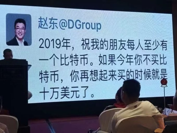 A conference screen displays a 2019 tweet by Zhao Dong (@DGroup) urging friends to buy at least one Bitcoin, warning that not buying now means paying $100,000 later. The image illustrates the post's point, highlighting how Dong's past $100,000 prediction mirrors the same hype-driven rhetoric used in current crypto price predictions.