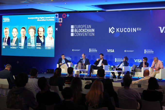 A panel discussion takes place at the European Blockchain Convention, with sponsors including KuCoin EU and Visa displayed on the stage backdrop above an audience.
