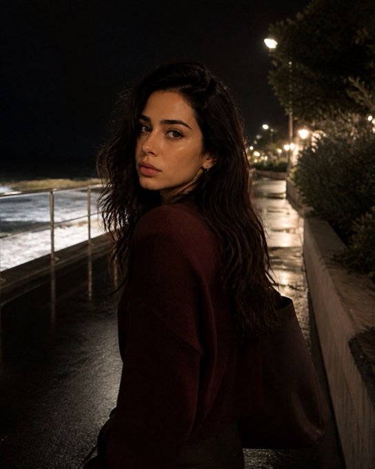 A young woman with long dark hair wearing a dark red sweater stands on a nighttime seaside walkway, looking over her shoulder toward the camera, with lights reflecting off the wet pavement.