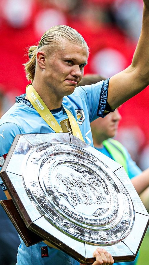 Erling Haaland, wearing a Manchester City kit and a "Winner" medal, holds the FA Community Shield shield after their victory. The image relates to the post by illustrating City's recent trophy success under their new manager, contextualizing the question of whether they can re-establish dominance over Arsenal in the upcoming season.