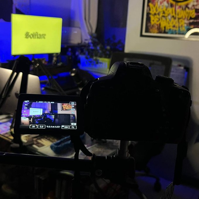 A camera on a tripod with its LCD screen displaying a live view of a streaming setup, with a monitor showing 'Solflare' and a framed poster on the wall.