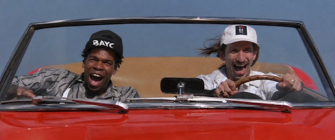 Two men laughing joyfully while riding in a red convertible car; the man on the left wears a black cap with 'BAYC' text, and the man on the right wears a white cap and grips the steering wheel.