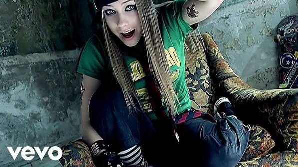 A screenshot from a music video showing a person with long blonde hair, wearing a green t-shirt and dark pants, sitting on a patterned couch with a Vevo logo in the corner.