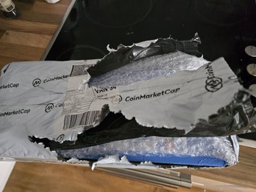 A torn and damaged shipping package from 'CoinMarketCap' lies on a wooden surface next to a stovetop, revealing bubble wrap and internal contents.