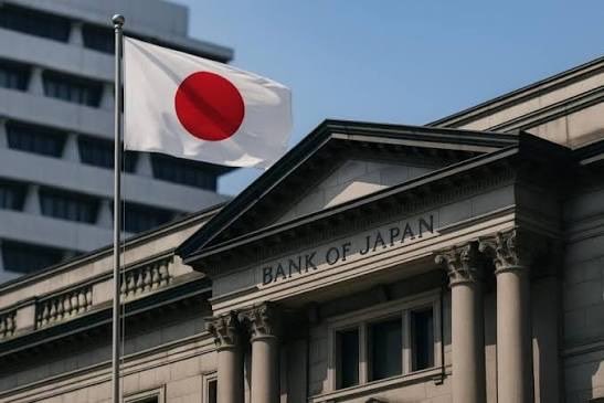 The image shows the facade of the Bank of Japan building with its "BANK OF JAPAN" sign prominently displayed on a classical columned structure, alongside a waving Japanese flag against a clear blue sky. This visual directly relates to the post's discussion of the upcoming Bank of Japan policy decision, highlighting the institution at the center of Friday's anticipated rate hike to 1.25%—a 31-year high that could impact yen-funded trades across global stocks and crypto markets.