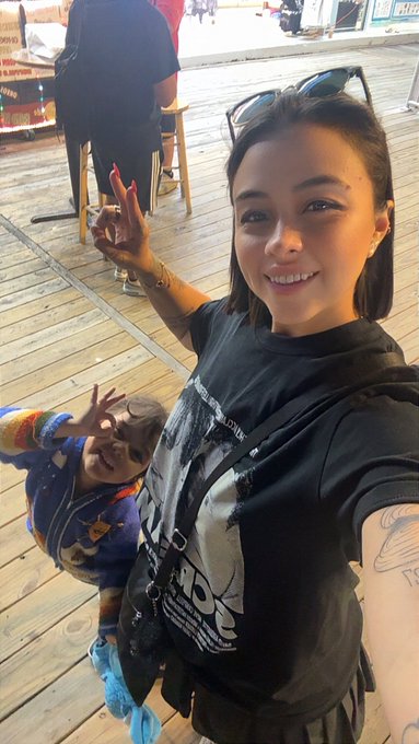 A smiling woman takes a selfie at what appears to be a theme park, with a child wearing a colorful jacket and sunglasses making a peace sign behind her.
