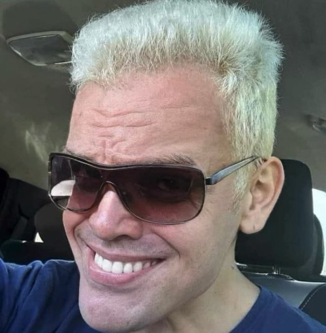 A smiling man with bleached blonde hair wearing sunglasses and a dark blue shirt, photographed inside a car.