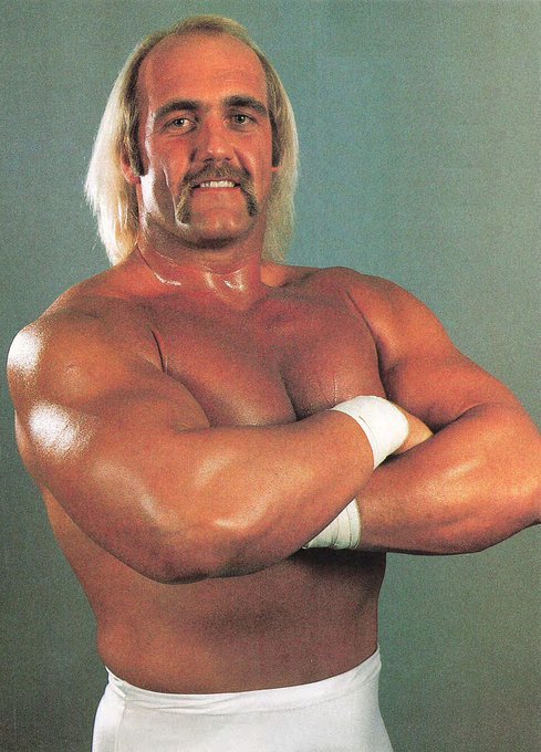 A muscular blonde wrestler with a mustache, tan skin, and crossed arms stands confidently against a teal background, wearing white wrist wraps and trunks — an iconic promotional portrait that visually matches the post's nostalgic reference to the legendary Hulk Hogan.