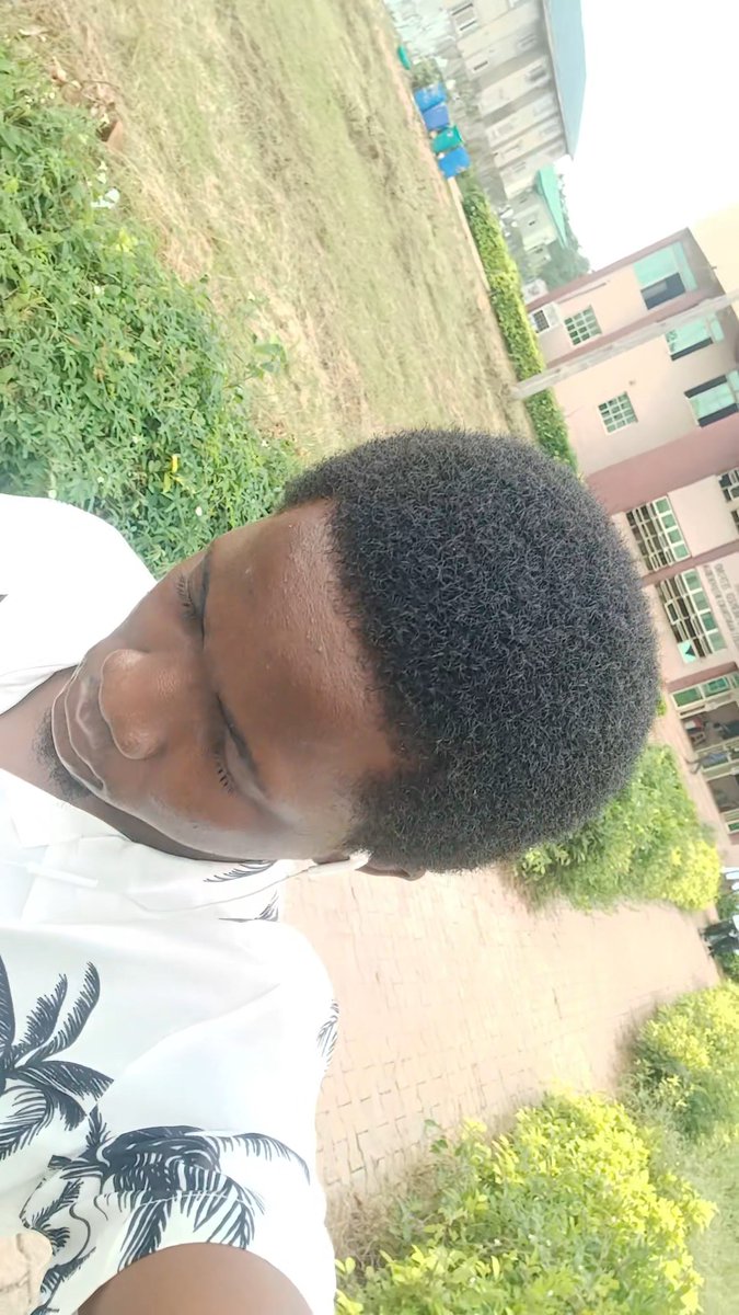 A sideways outdoor selfie shows a man in a white shirt with a dark leaf pattern standing near grass, shrubs, a brick path, and apartment buildings.