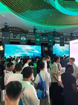 A crowded indoor event hall shows people gathered beneath green ceiling lights, with large screens displaying “Invest in your passion / Live out of passion” and a Bose sign visible.