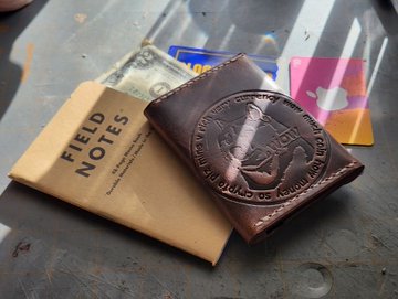 A brown leather Field Notes notebook and a brown leather wallet embossed with a logo, resting next to some currency on a scratched metal surface.