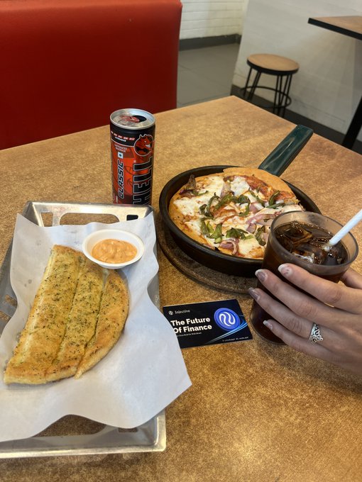 A meal setup on a table with garlic bread, a small bowl of orange sauce, an energy drink can, a skillet pizza, and a hand holding an iced soda next to an 'Intuit' business card.