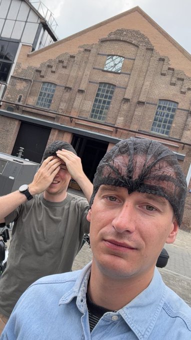 A selfie of two young men wearing black mesh shower caps (bubble cap style) standing in front of a large brick industrial building.