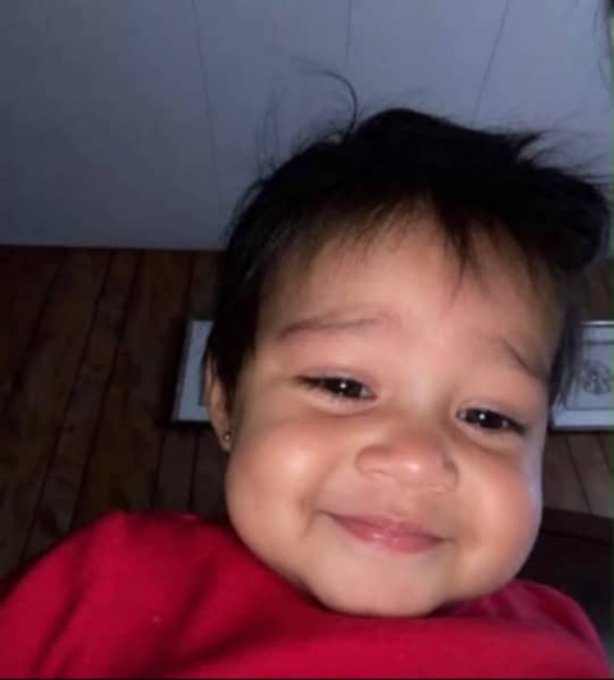 A close-up smiling selfie of a young child with short dark hair wearing a red top against a white background.