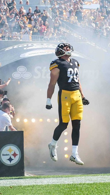 A Pittsburgh Steelers football player wearing jersey number 90 leaps into the air through a cloud of fog during a stadium entrance, with the Steelers logo visible on the wall behind him.