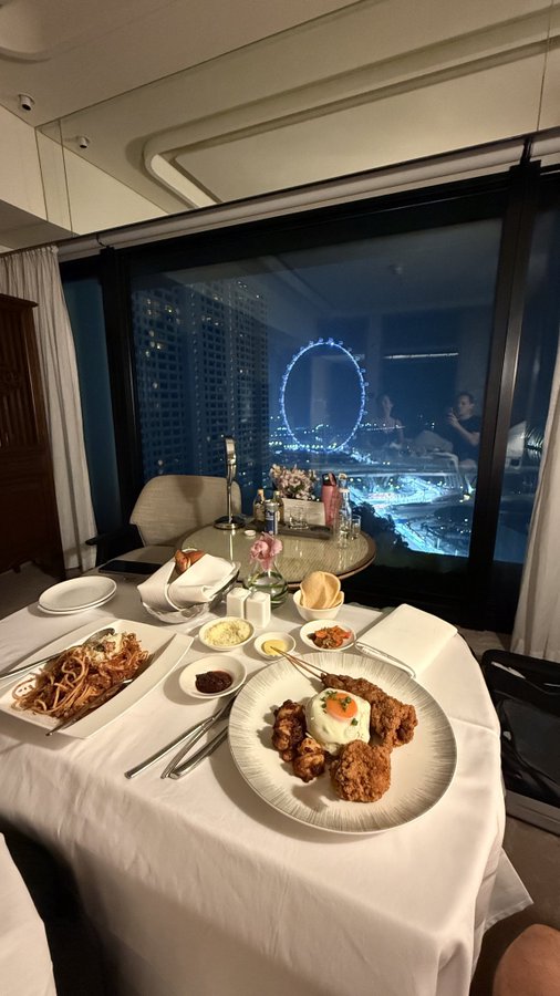 A couple enjoys a gourmet dinner of fried cutlets, pasta, and appetizers at an upscale table-for-two set against a stunning nighttime view of Singapore's Marina Bay Sands and the Singapore Flyer Ferris wheel. The scene captures a memorable romantic evening in Singapore during their Token2049 and F1 trip.