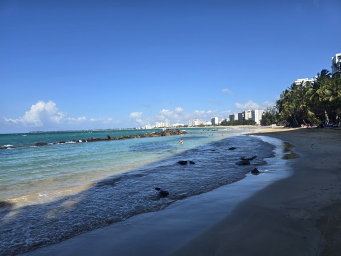 A scenic view of a tropical beach with turquoise water, gentle waves, palm trees on the right, and high-rise buildings in the distance under a bright blue sky with scattered clouds.