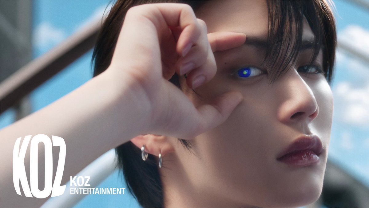 The image shows a young person with dark hair holding up their hand to reveal a striking glowing blue eye, with the "KOZ Entertainment" logo visible in the corner. The caption text overlay appears alongside what seems to be a teaser or promotional still, with the fan's message expressing excitement for new music releases.
