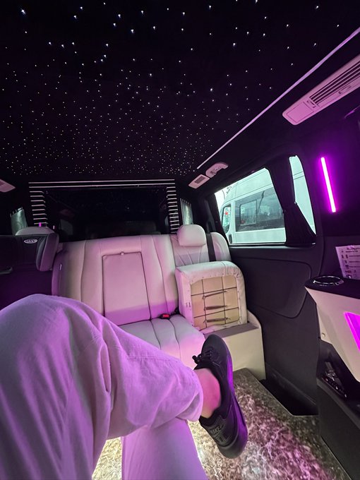 An interior view of a luxury van featuring a starlight ceiling with small LED lights, cream-colored leather captain's chairs, and purple ambient lighting, with a person's legs stretched out in the foreground.