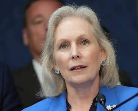 A close-up portrait of an older woman with grey hair wearing a blue jacket, appearing to be at a press conference or public event.