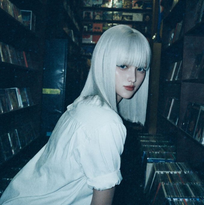 A young woman with platinum blonde hair in a bob cut poses in a white blouse, standing in what looks like a record store aisle with shelves of media behind her.