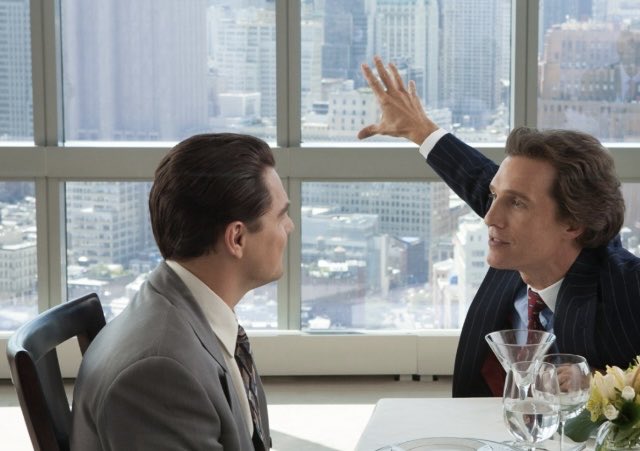 Two men in suits sit at a table in a high-rise office with a city view; one man raises his hand dramatically as if making a point.