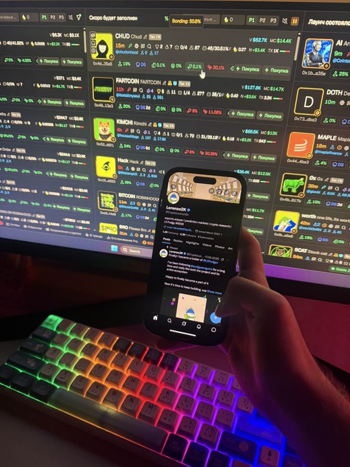 A person uses a smartphone in front of a monitor showing a crypto meme coin dashboard and a backlit RGB keyboard.