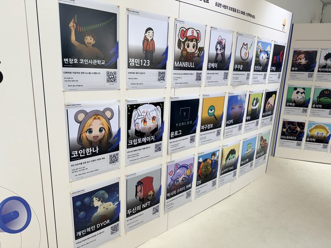 A display board at a Korean event showcases various NFT character collections with Korean names like 번창훈, 챔피123, MANBULL, 코인헌터, and 토신의 NFT, each accompanied by a QR code.