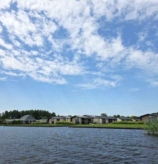 A scenic landscape photograph of modern waterfront houses or vacation homes along a calm lake, with a beautiful blue sky filled with scattered white clouds above.