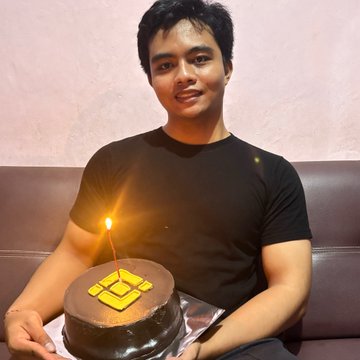 The image features a man sitting on a couch, holding up a chocolate cake with a lit candle on top. The cake has a yellow diamond design on its side. The man appears to be celebrating his birthday or possibly another special occasion. The background of the photo is blurred and does not provide any additional information about the event taking place in the image.